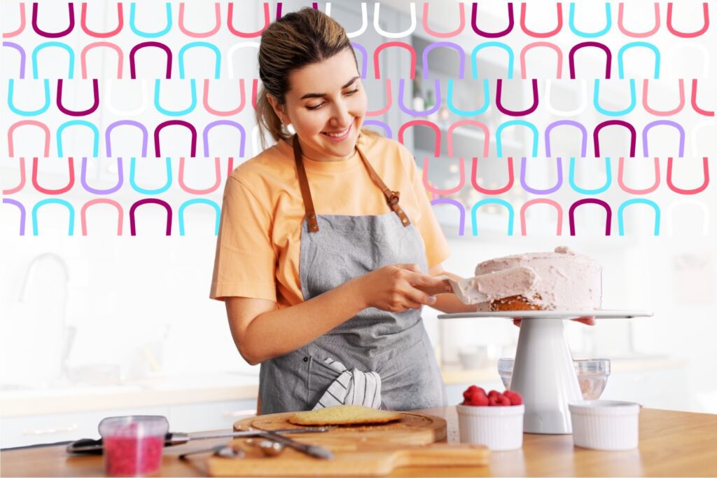 Microempreendedora confeiteira sorridente, de avental cinza, cobrindo um bolo rosa na cozinha; imagem usada para ilustrar os benefícios de um plano odontológico para MEI.